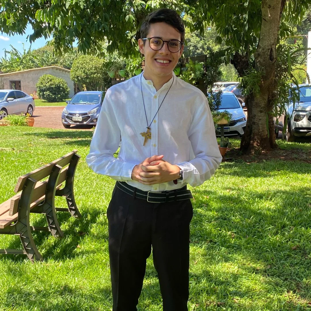 Retrato de Pedro Henrique Godoy Costa sorrindo, de camisa branca e cruz no peito, em um jardim